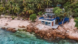 A beachfront property with a small building, a rocky shoreline, and palm trees. - P.D. Beach Resort (Koh Tao)