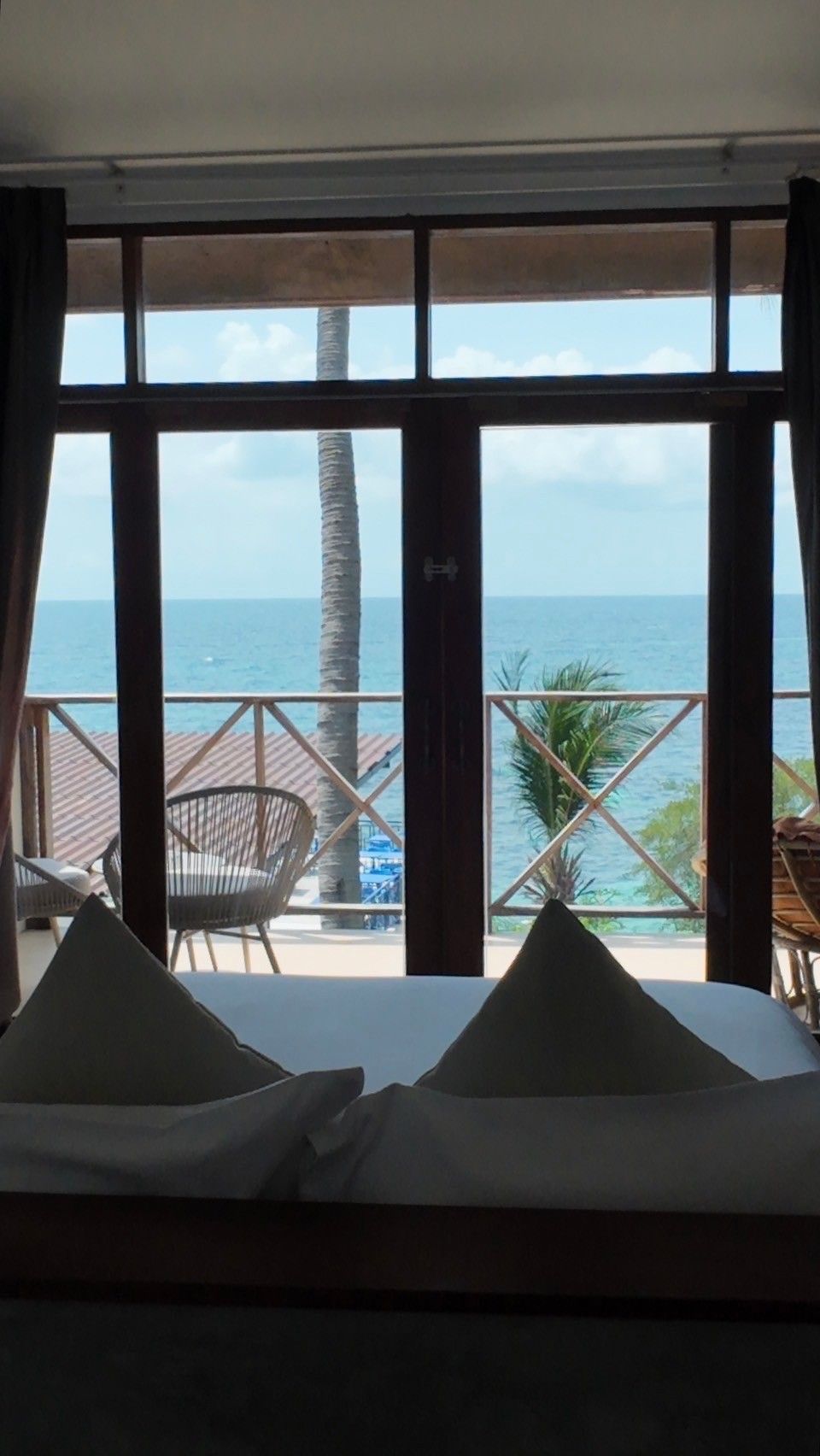 A room with a large window overlooking a beach with palm trees and a wooden deck.