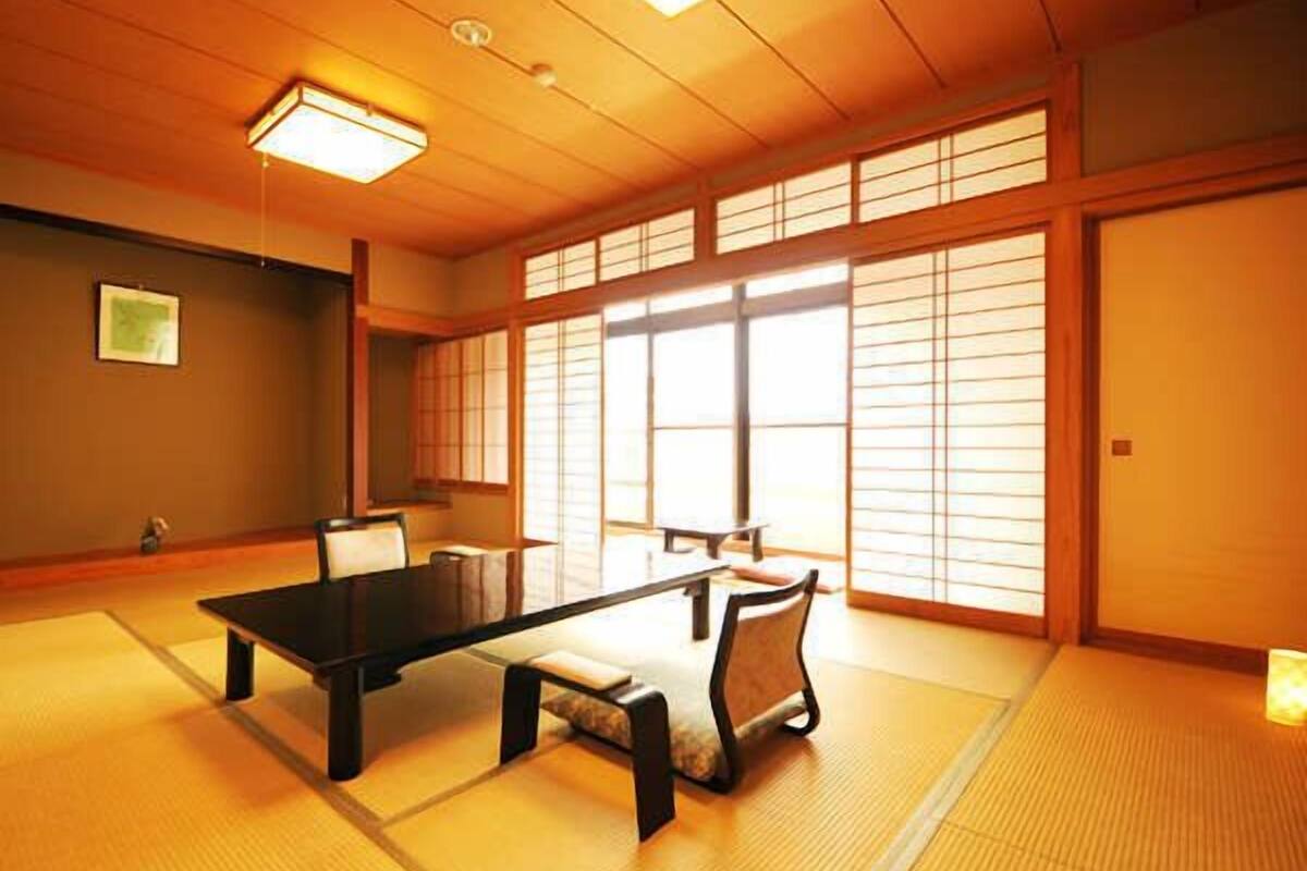 A tatami mat room with a low table and chairs, large windows with shoji screens, and a ceiling light.