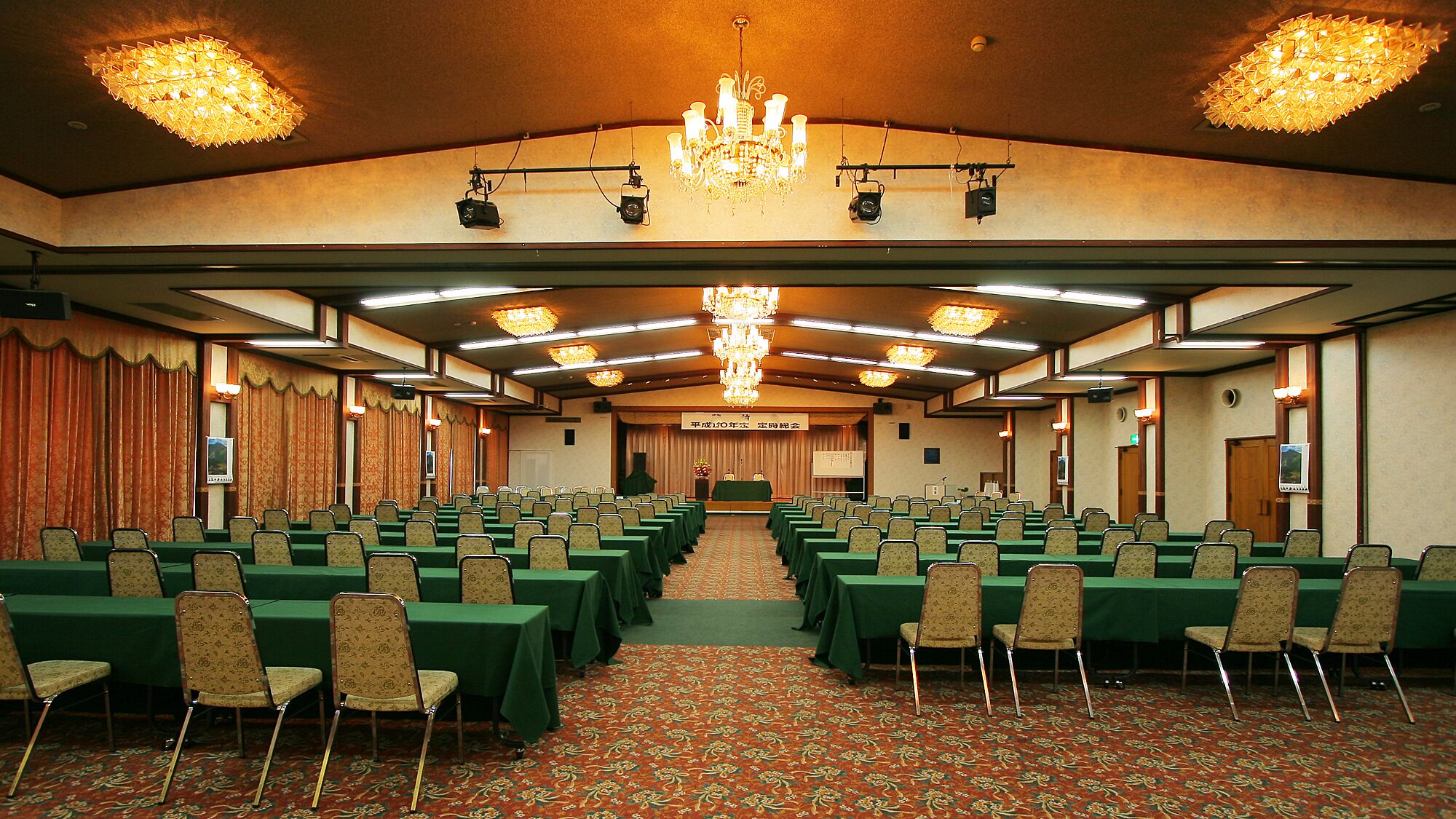 A conference room with rows of tables and chairs, a chandelier, and a stage with a banner.
