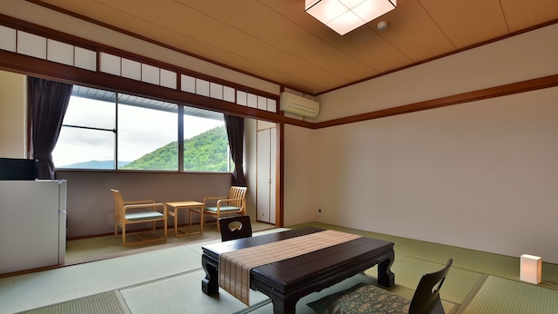 Une pièce de style japonais traditionnel, avec un sol recouvert de tatamis, une table basse et un banc, deux chaises, un réfrigérateur et une fenêtre donnant sur des collines verdoyantes.