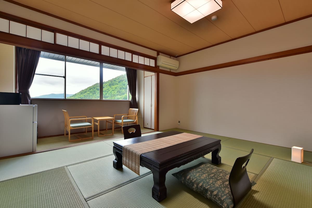 Ein traditioneller japanischer Raum mit Tatami-Böden, einem niedrigen Tisch mit einer Bank, zwei Stühlen, einem Kühlschrank und einem Fenster mit Blick auf grüne Hügel.