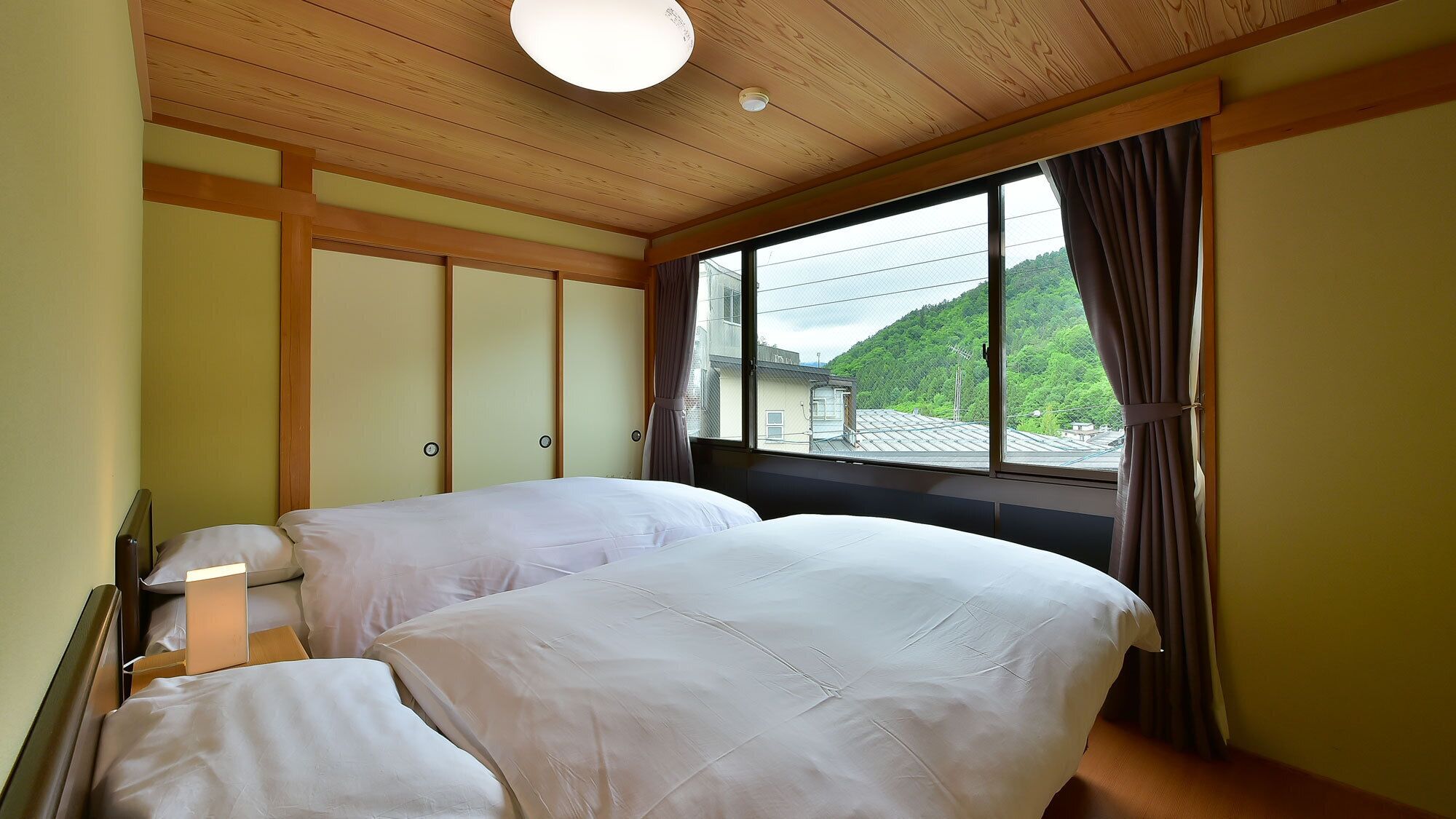 Ein Zimmer mit zwei Betten, einem großen Fenster mit Blick auf Grünflächen, Holzboden und einer Lampe.