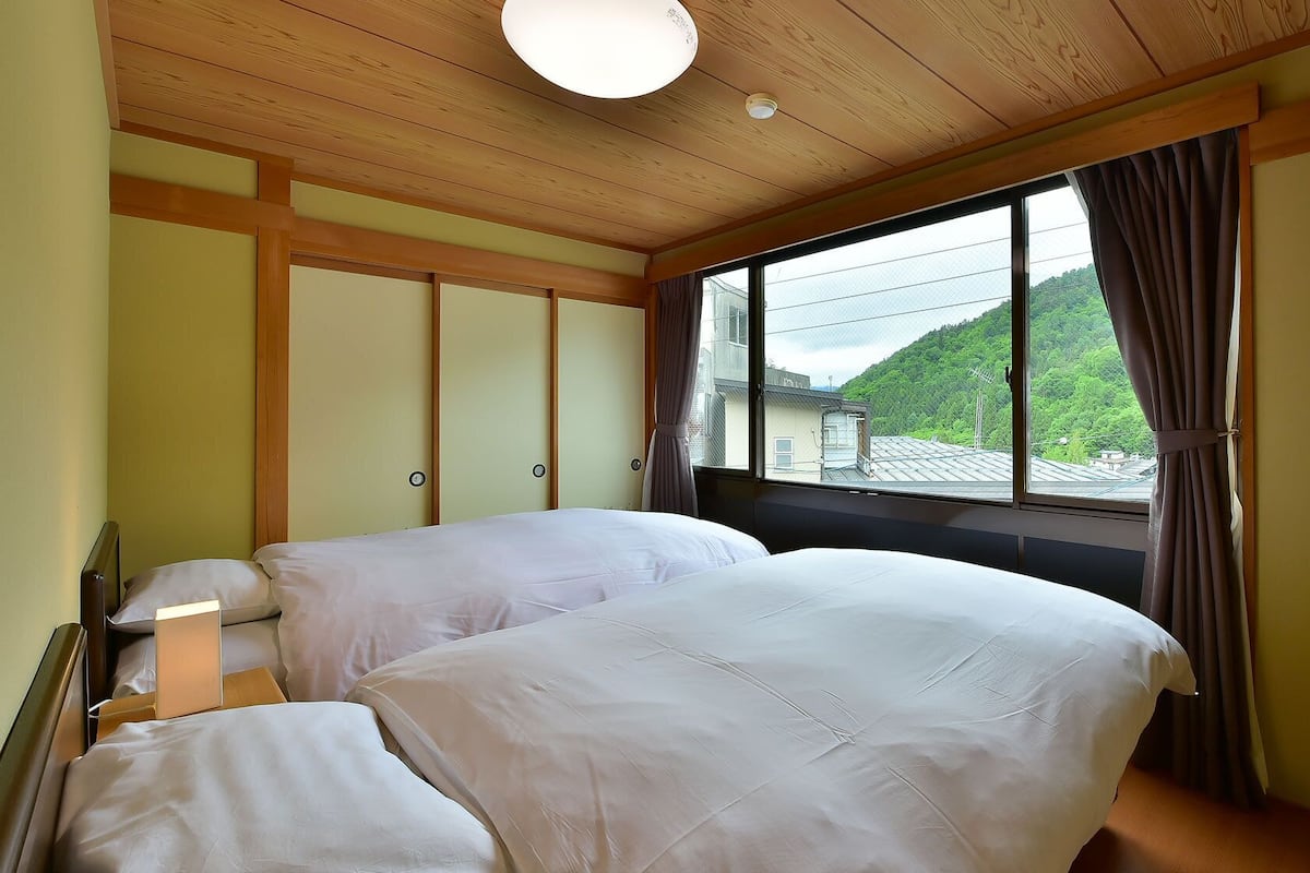 Ein Zimmer mit zwei Betten, einem großen Fenster mit Blick auf Grünflächen, Holzboden und einer Lampe.