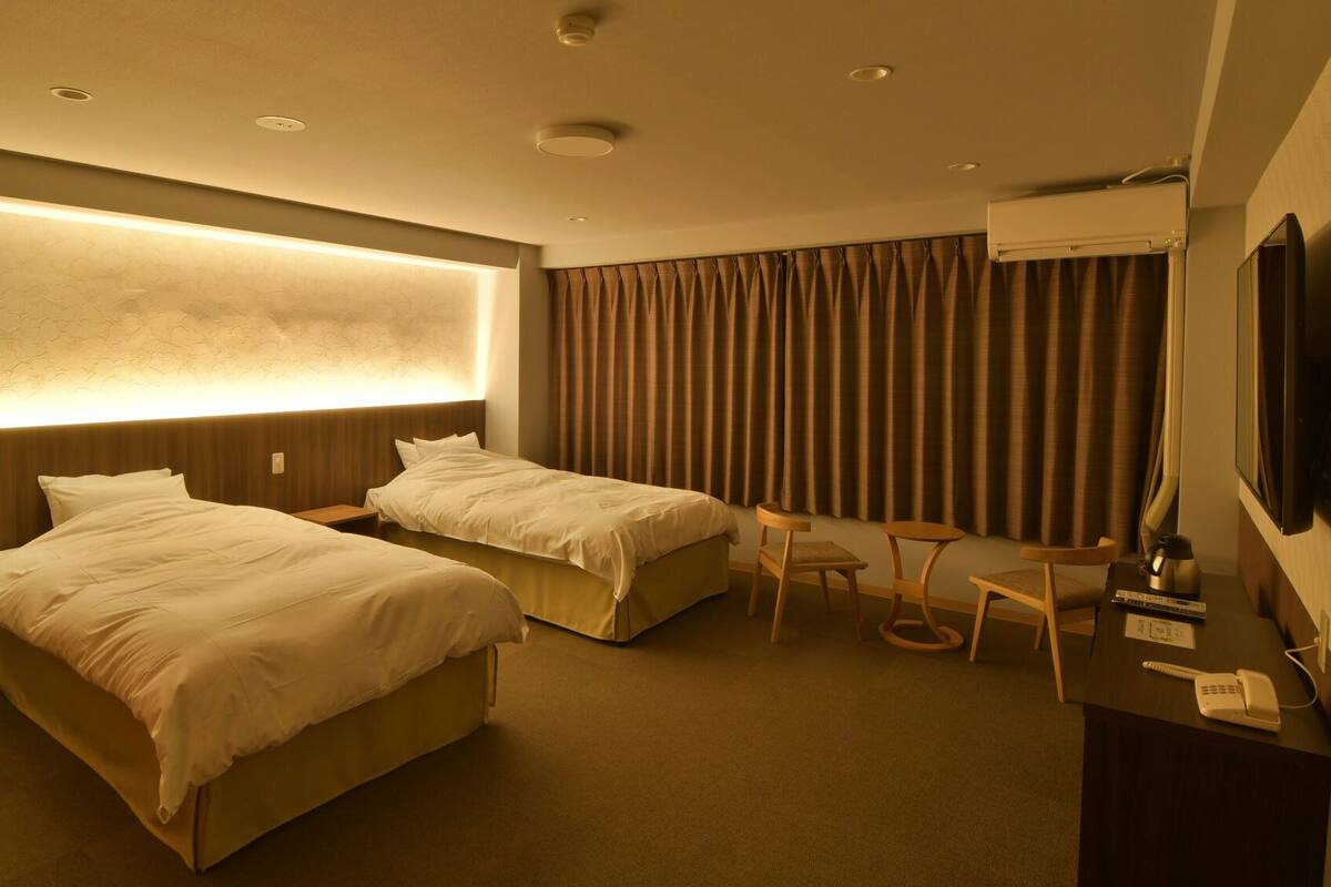 Ein Hotelzimmer mit zwei Betten, einem Schreibtisch, einem Sessel, einem Fernseher und einer Klimaanlage.