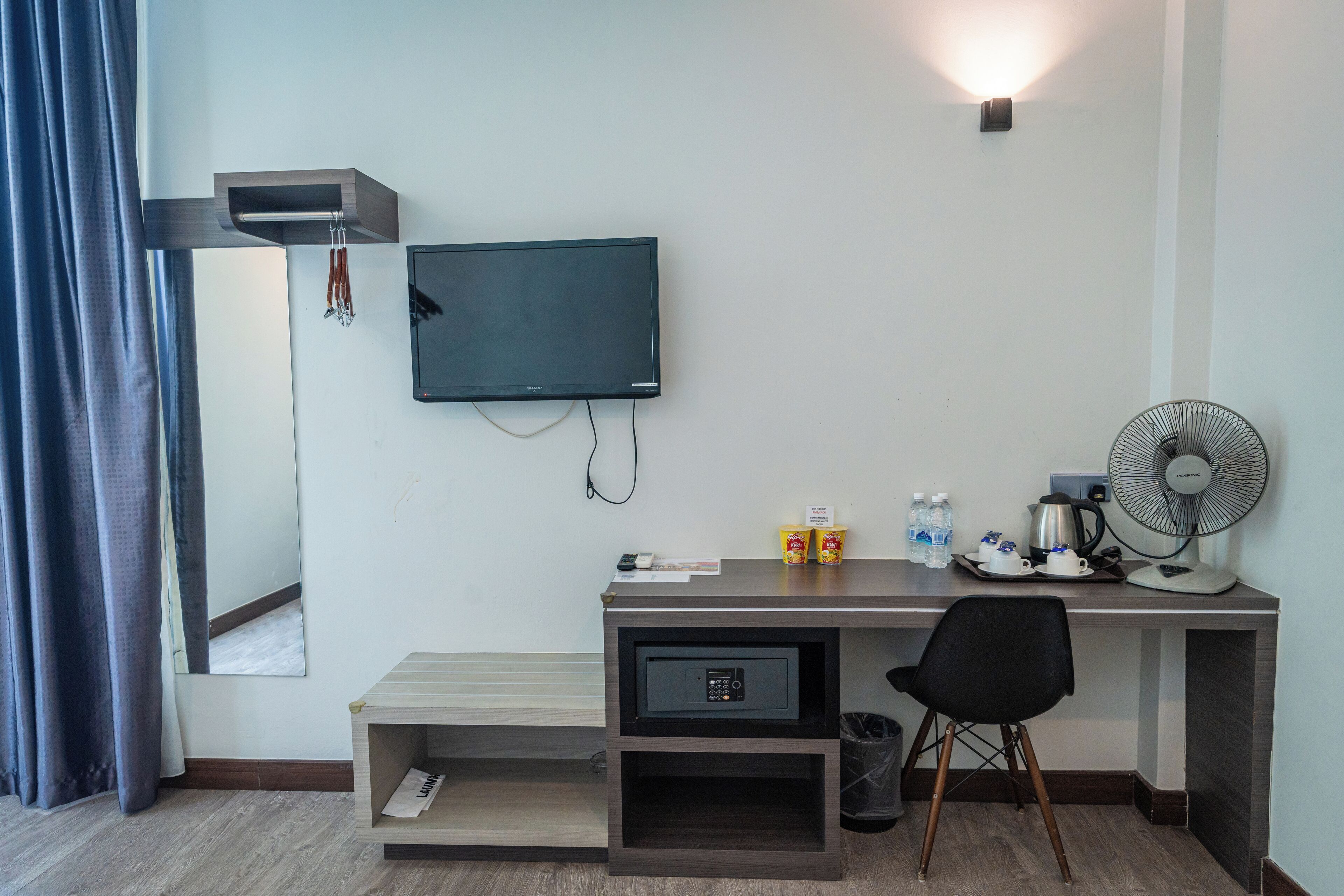 A hotel room with a desk, a television mounted on the wall, a fan, a trash bin, and a window with curtains.