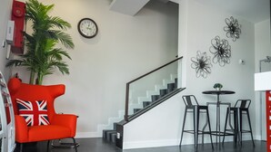 A modern living room with a staircase, a red chair with a Union Jack cushion, a potted plant, and a wall clock. - Indra Hotel (Ipoh)