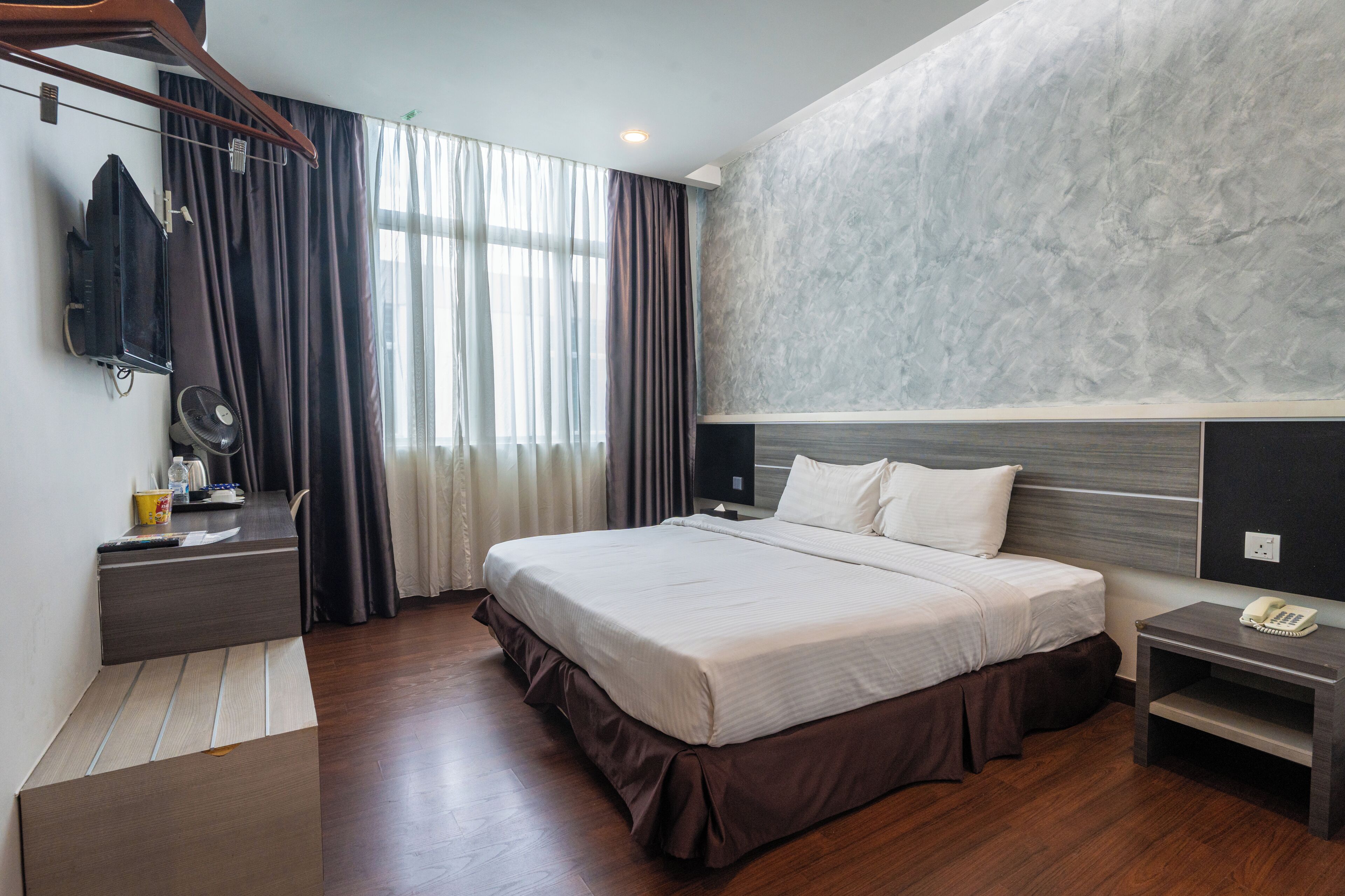 Een moderne hotelkamer met een groot bed, een nachtkastje met telefoon, een bureau met ventilator en uitzicht op het buitengebied door het raam.