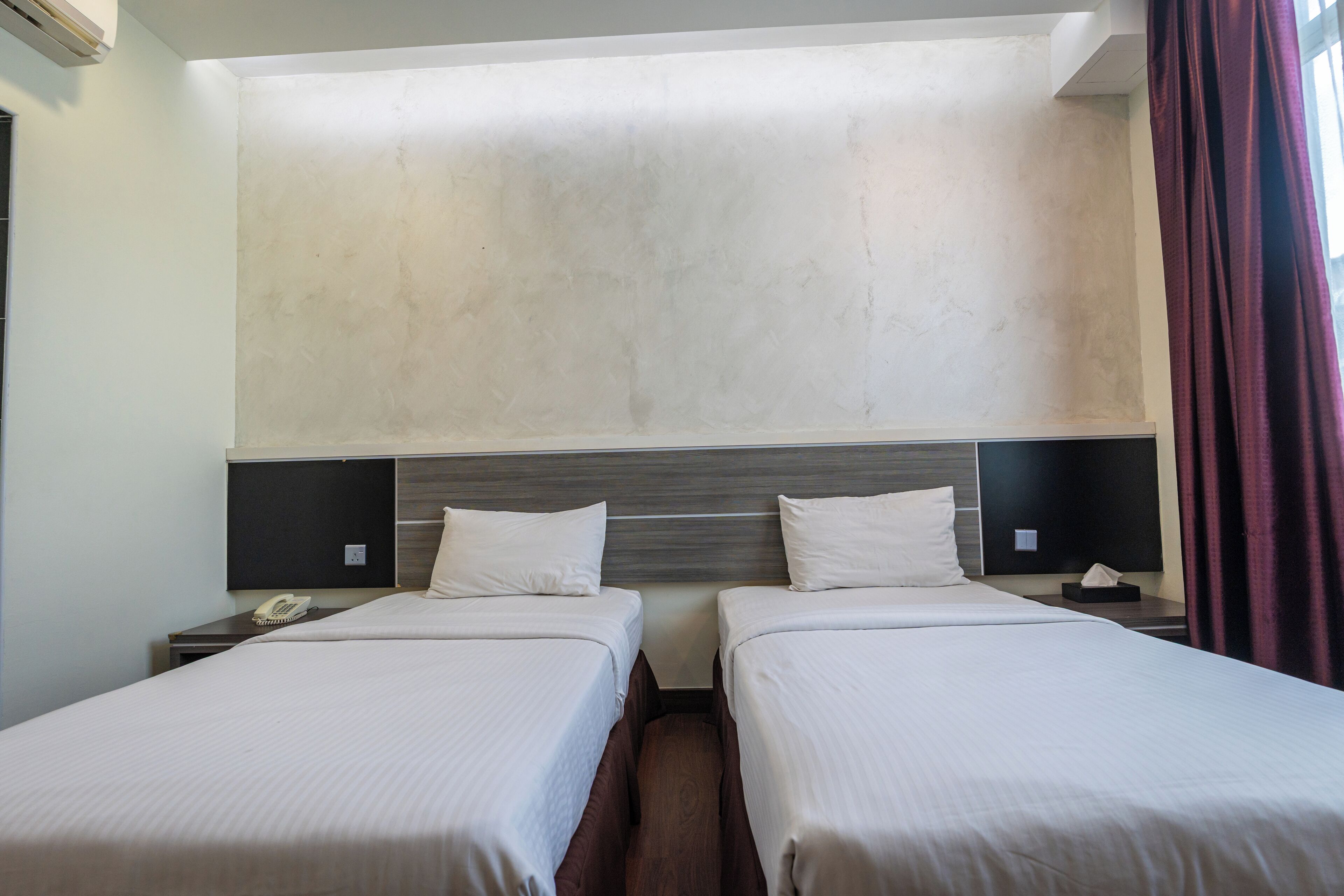 A hotel room with two beds, a large window with curtains, a wall-mounted air conditioner, and a bedside table with a phone.