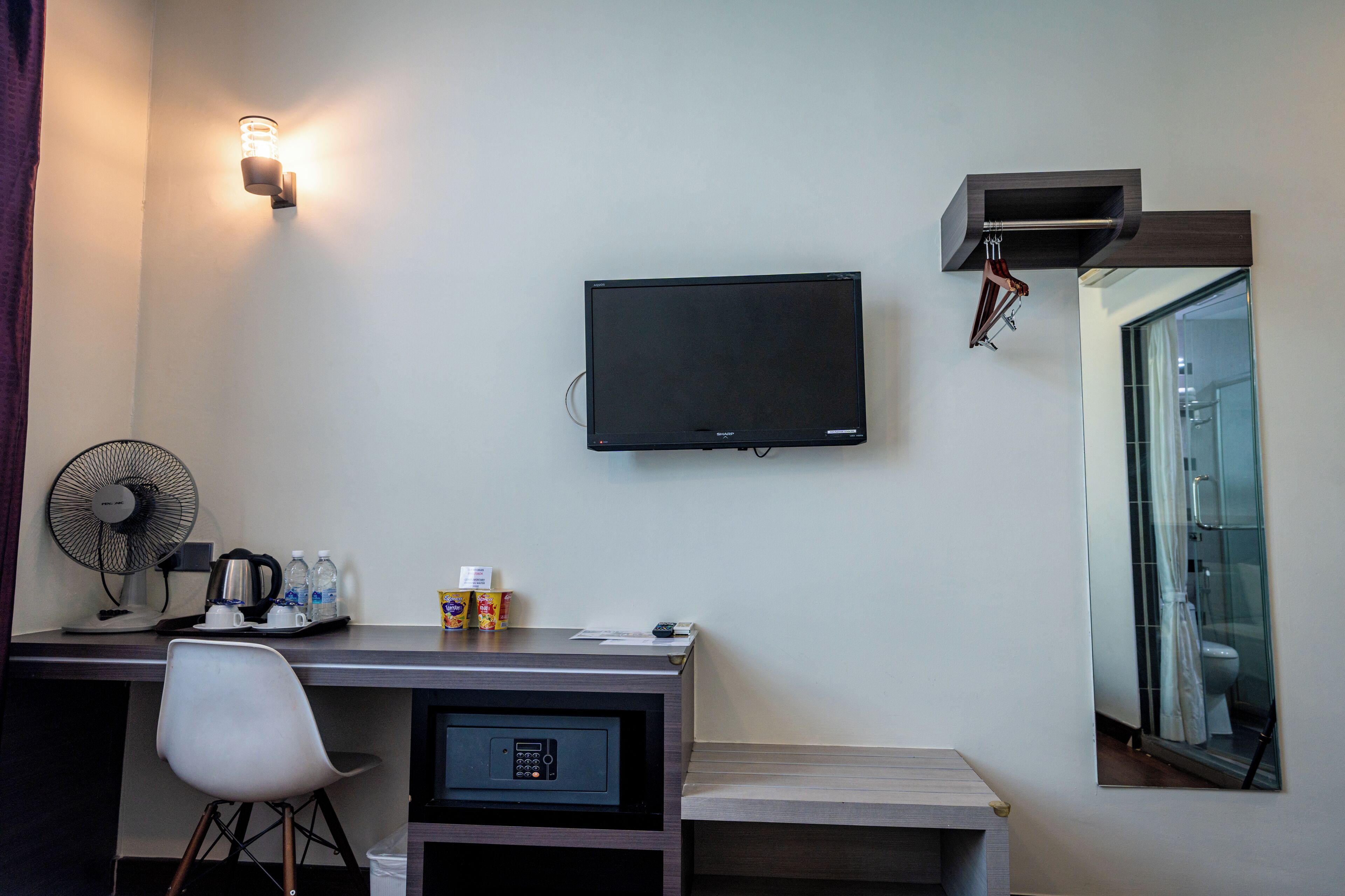 A hotel room with a desk, chair, flat-screen TV, fan, and a mirror reflecting the bathroom.
