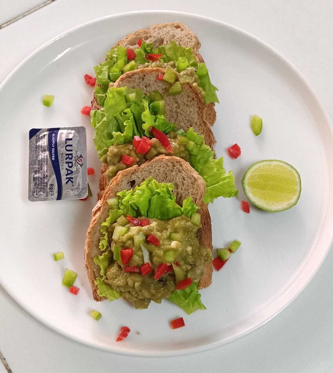 Un plato con rebanadas de pan cubiertas de guacamole y pimientos rojos picados, acompañado de un gajo de lima y una porción de mantequilla Lurpak.