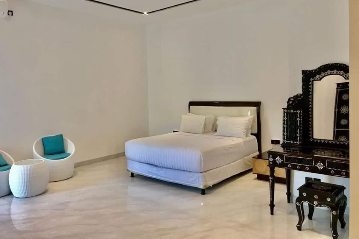 A modern bedroom with a bed, two white chairs, a wooden dresser, and a large mirror.