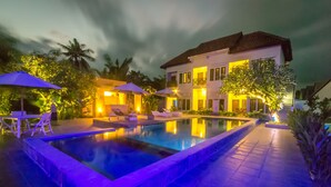 Una piscina con sillas de descanso y sombrillas, rodeada por un edificio blanco con balcones, palmeras y una terraza con baldosas.
