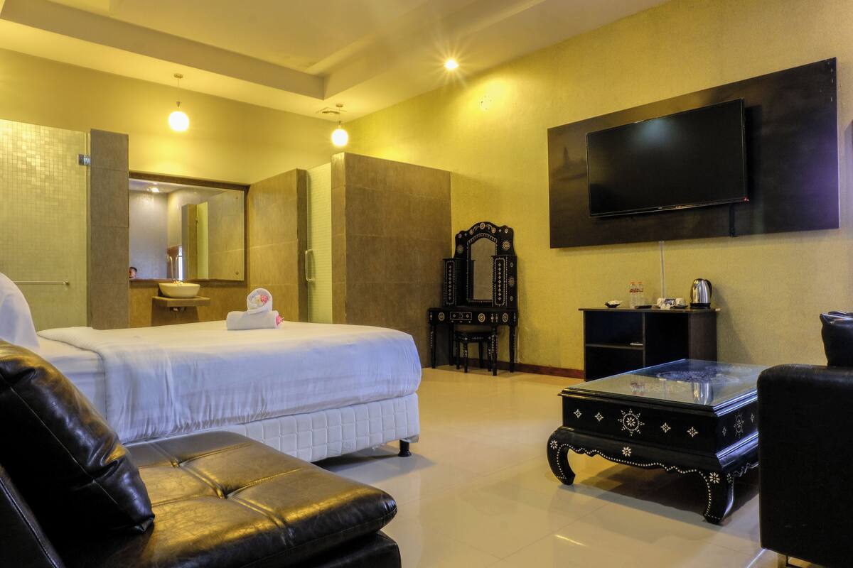 A hotel room with a bed, a black leather sofa, a glass coffee table, a flat-screen TV, and a decorative mirror.