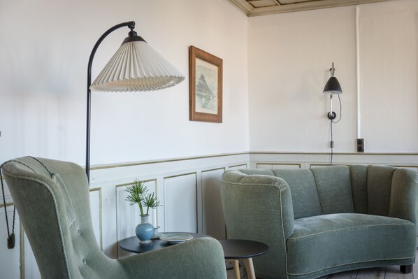 Una habitación con un sillón verde, un jarrón azul con una planta, una mesita redonda y dos lámparas fijadas en la pared.