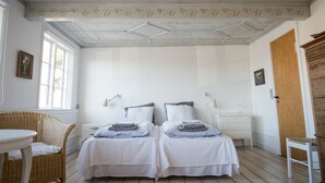 A room with two beds, a wicker chair, a small table, and a wooden door. - Pension Holmegaard (Allinge)