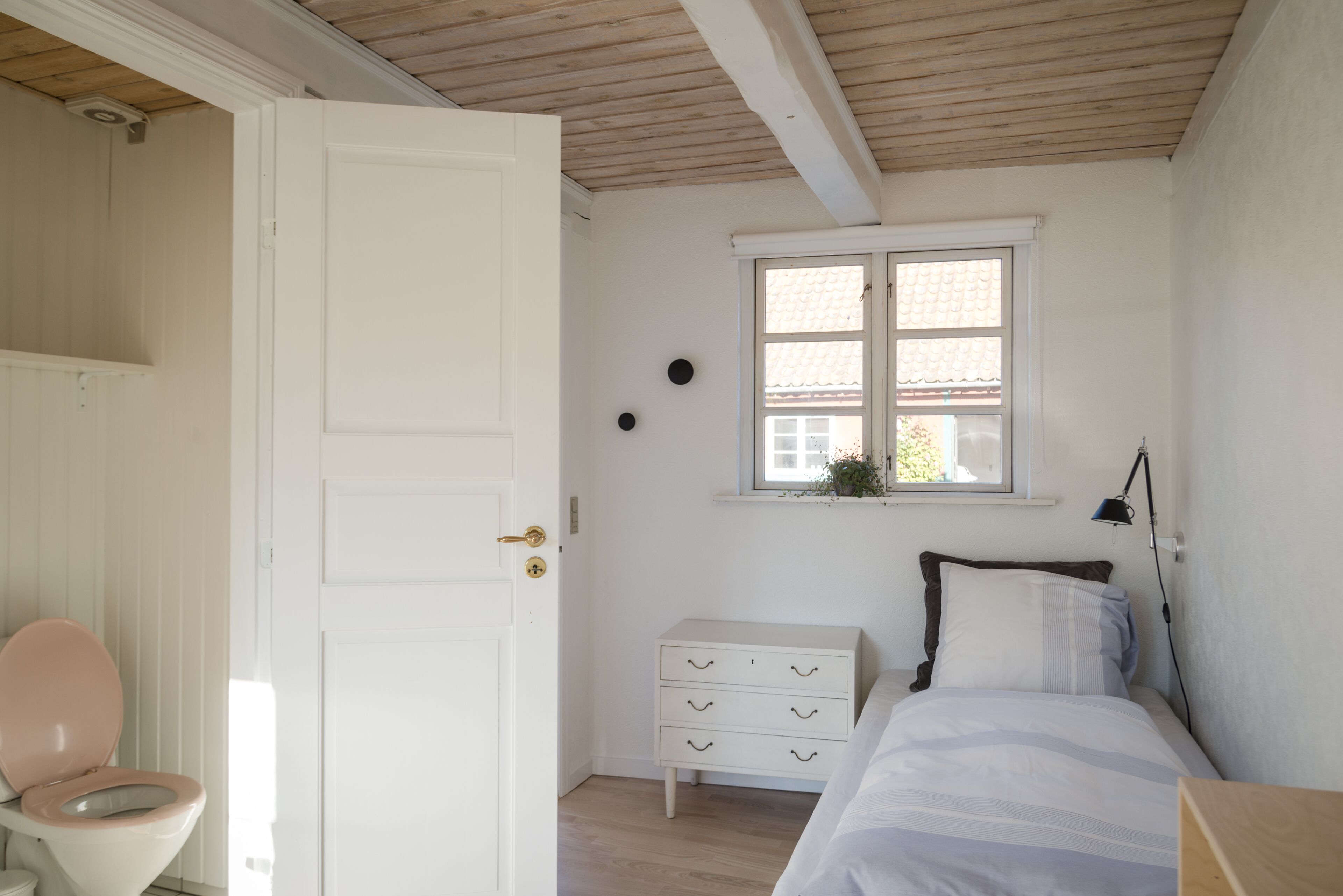 A bedroom with a white door, a bedside table with a lamp, a bed with white bedding, and a window with a view of a house.