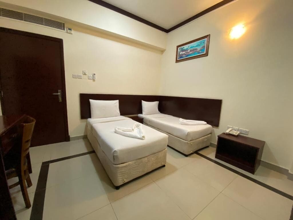 A hotel room with two beds, a desk, a chair, a bedside table, and a wall-mounted light.