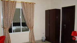 A bedroom with a bed, a wardrobe, a window with curtains, a red chair, and a trash bin. - Savoy Grand Hotel Apartments (Muscat)