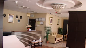 A reception area with a reception desk, a red patterned rug, a sofa, and a potted plant. - Savoy Grand Hotel Apartments (Muscat)