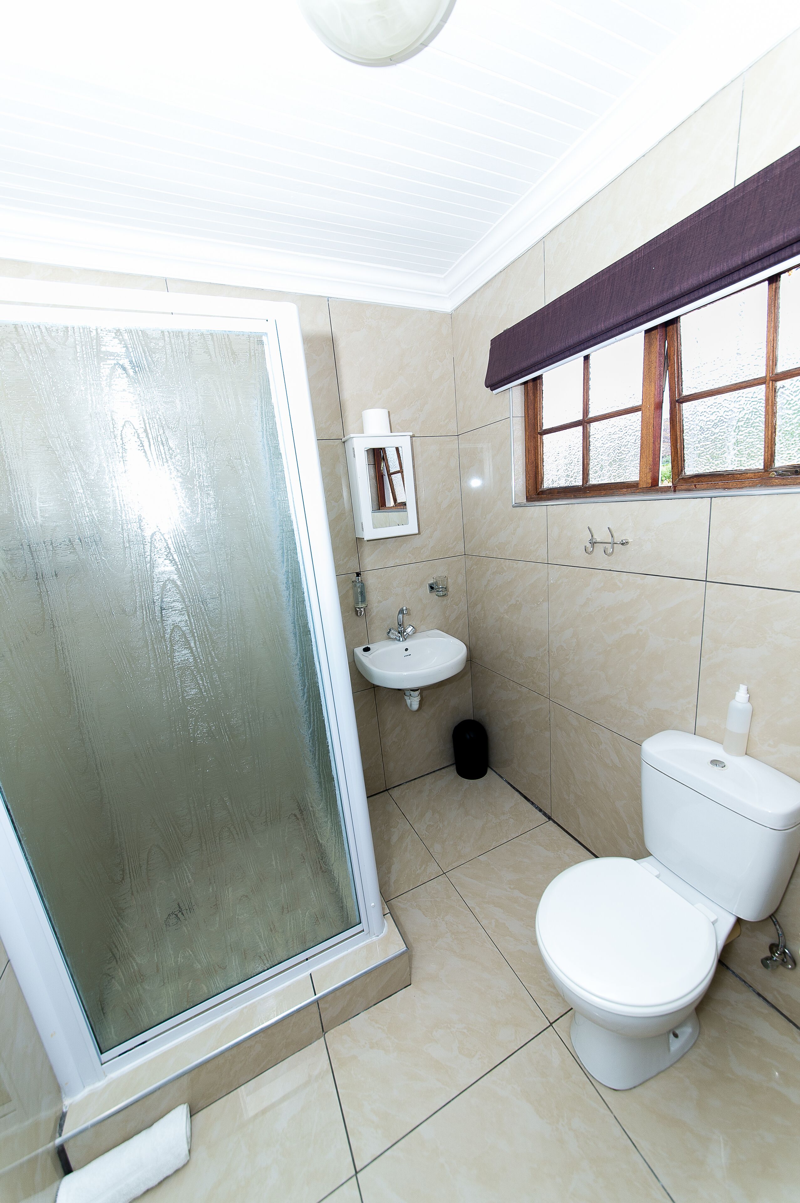 Een badkamer met een glazen douchedeur, een wastafel, een toilet en een raam met een bruin gordijn.