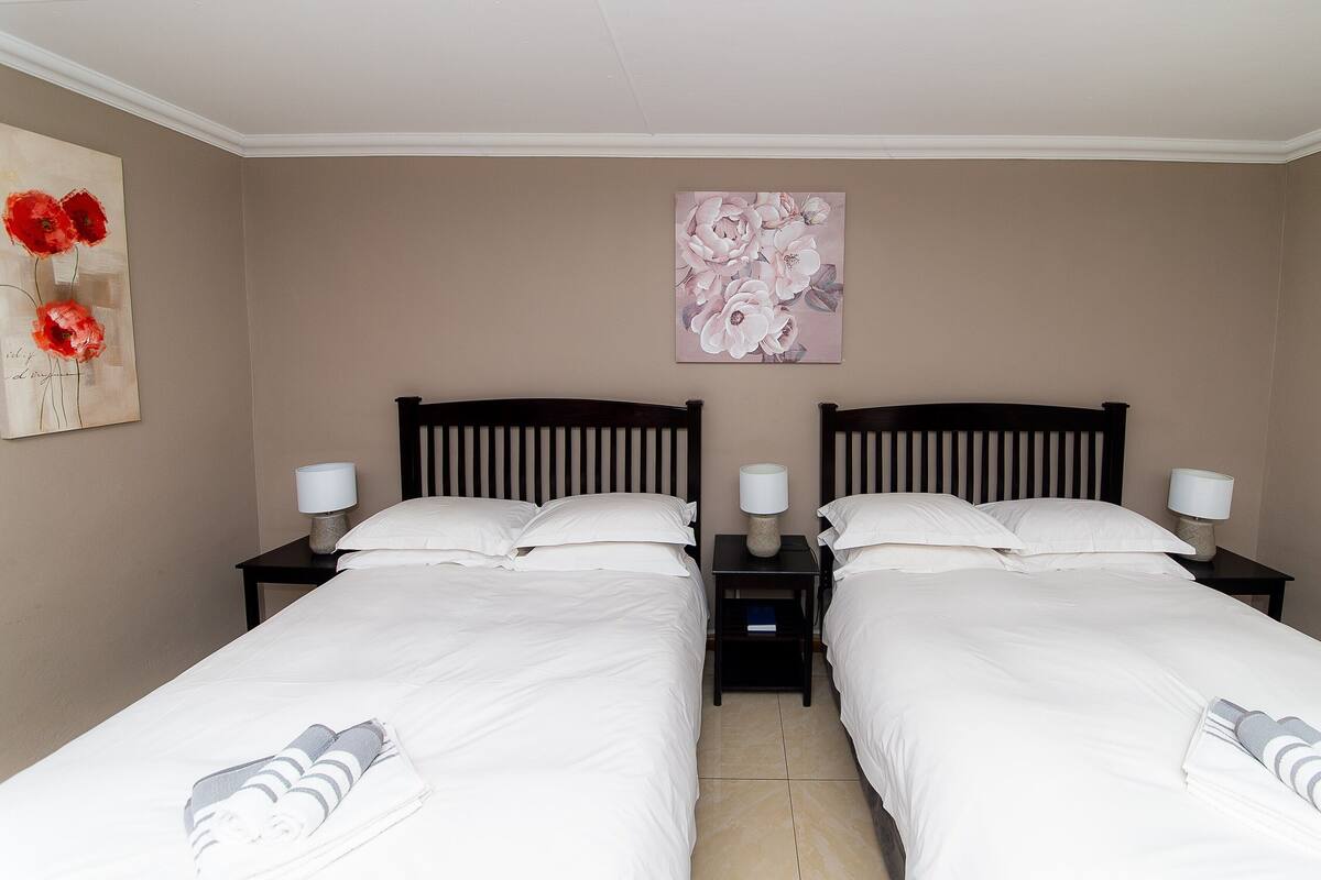 Ein Hotelzimmer mit zwei Einzelbetten, jeweils mit einem Nachttisch und einer Lampe. Darüber hängt ein Blumenbild an der Wand.