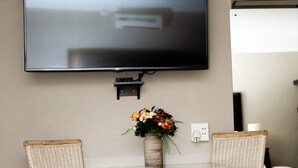 Ein Wohnzimmer mit einem an der Wand befestigten Flachbildfernseher, einem kleinen Tisch mit Blumen in einer Vase und zwei Korbstühlen.