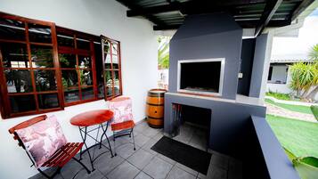 A patio area with a grill, two chairs, a table, and a window with a view of greenery.