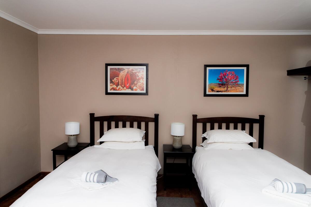 Ein Hotelzimmer mit zwei Einzelbetten, jeweils mit Nachttisch und Lampe. An der Wand hängen zwei gerahmte Bilder.
