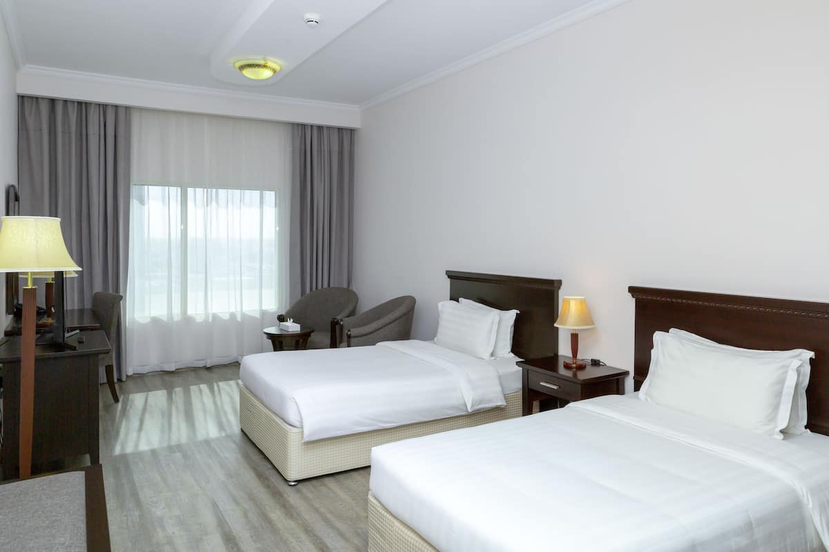 A hotel room with two beds, a desk, a chair, and a lamp.