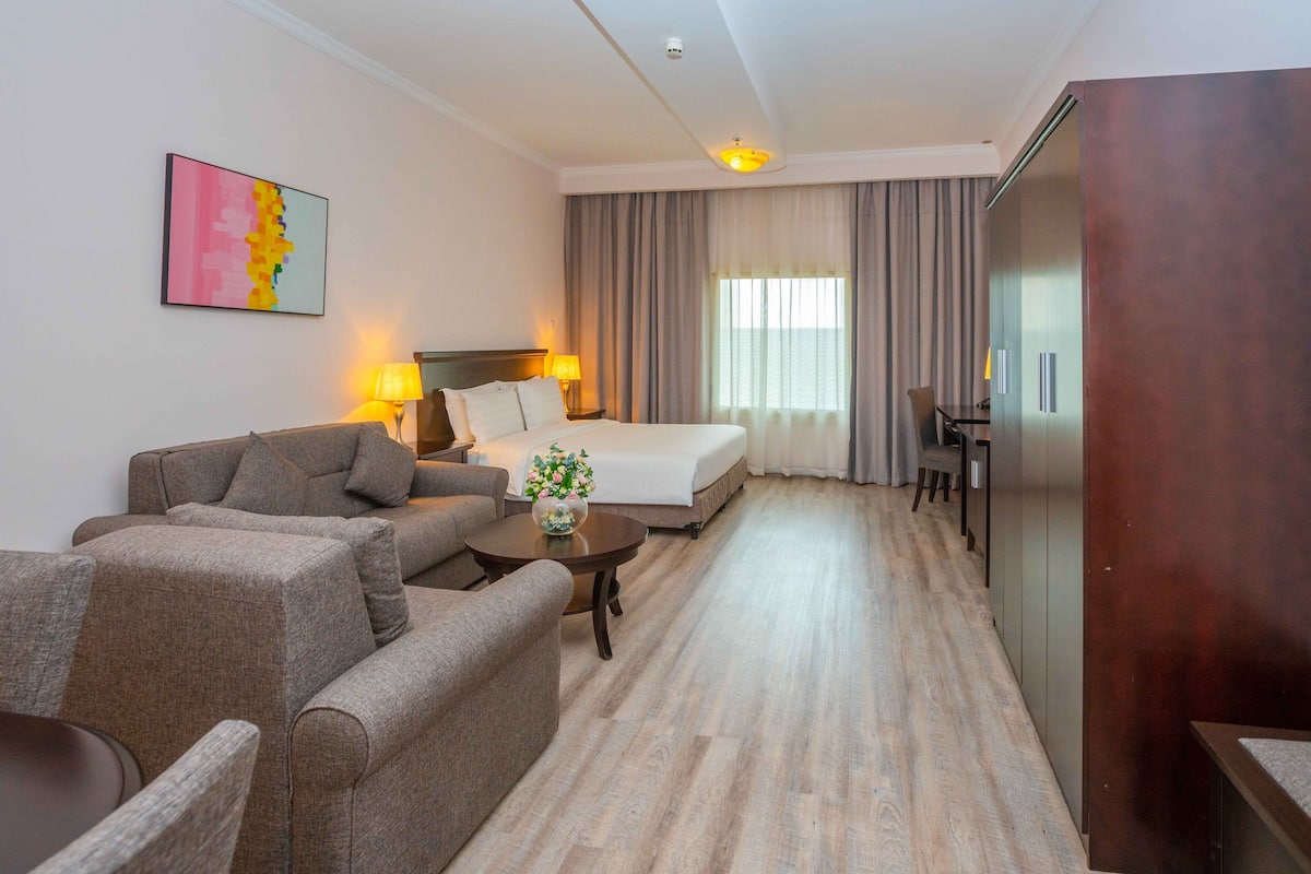A hotel room with a bed, two sofas, a coffee table with flowers, a desk, and a large window with curtains.