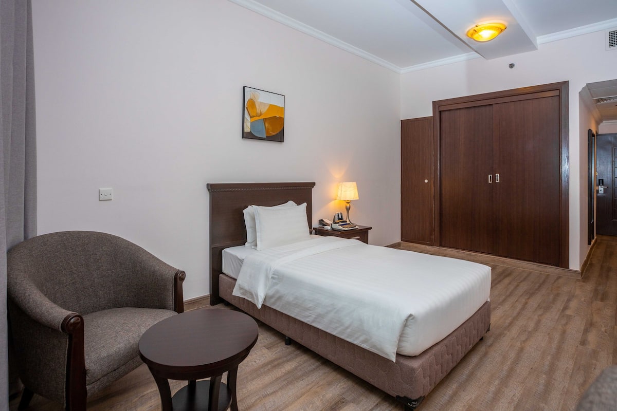 A hotel room with a bed, a chair, a small table, a lamp, and a wardrobe.