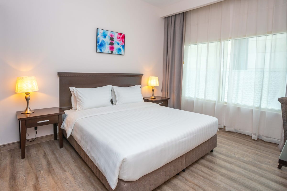 A hotel room with a large bed, two bedside lamps, a nightstand, a chair, a window with curtains, and a colorful abstract painting on the wall.