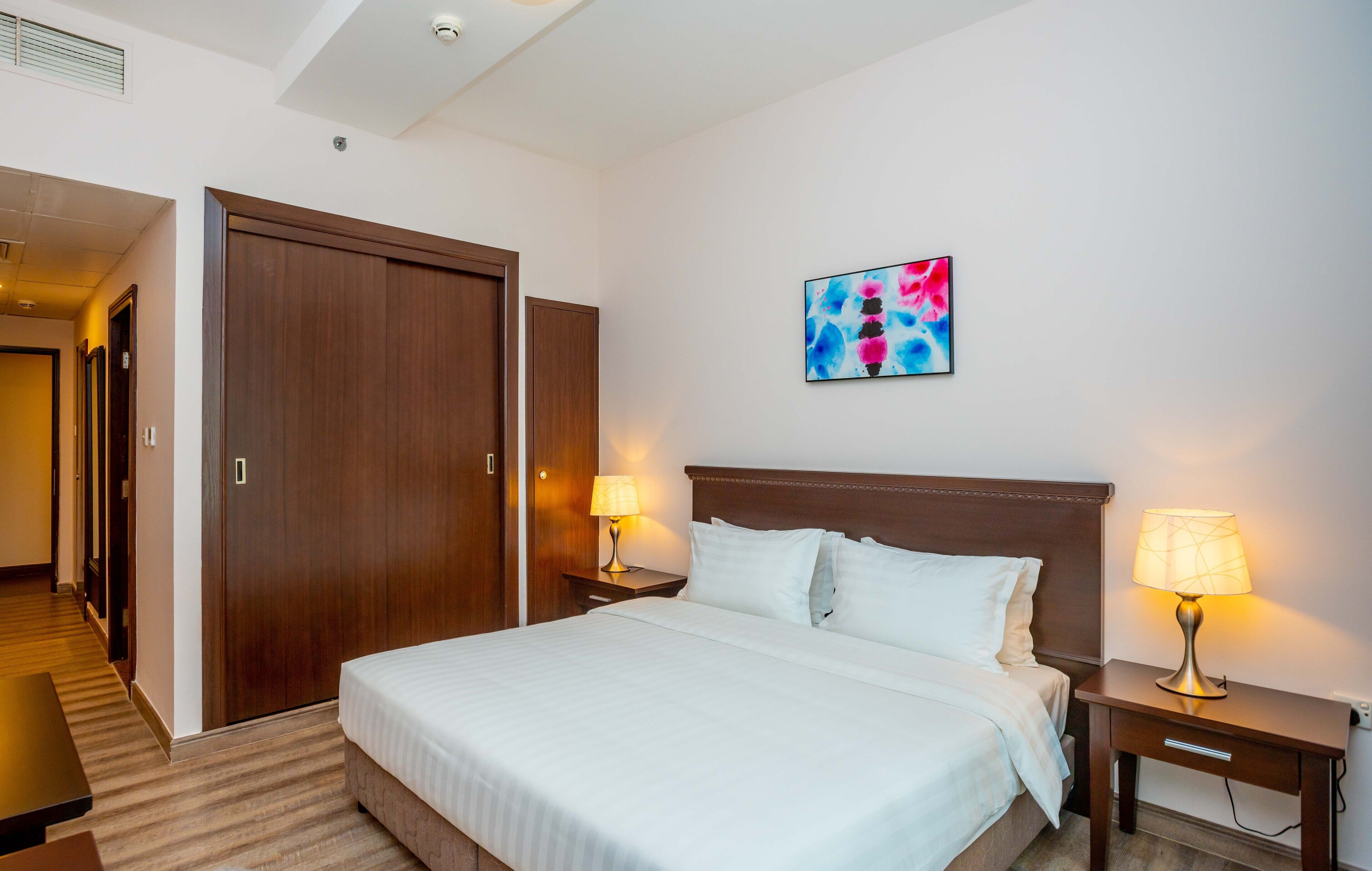 Camera d'albergo con un letto grande, due comodini con lampade, un armadio in legno e un quadro astratto dai colori vivaci appeso al muro.