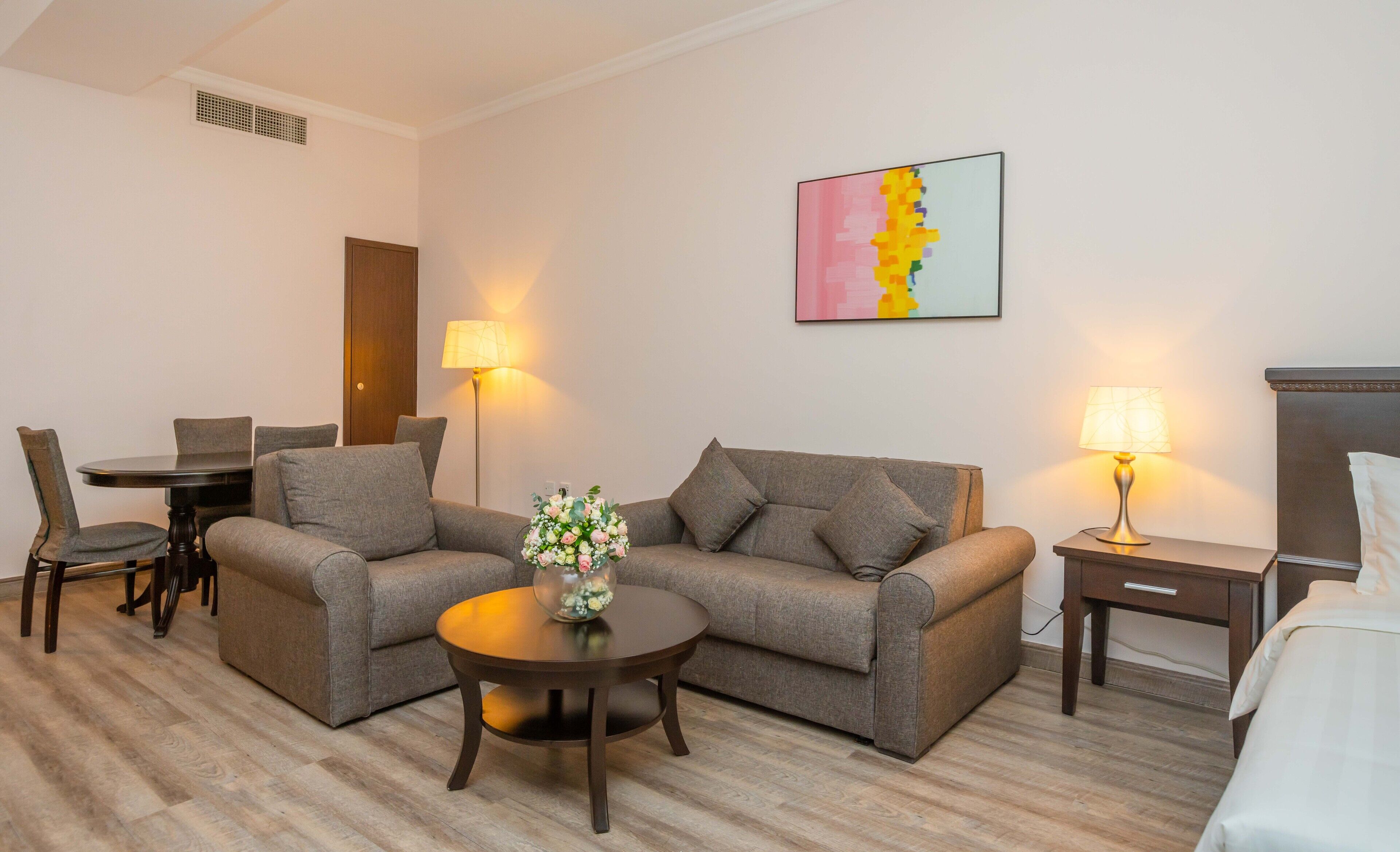 Una habitación de hotel con un conjunto de sofá, una mesa de comedor con sillas, una mesita redonda con un jarrón, una mesita de noche con una lámpara y una cama.