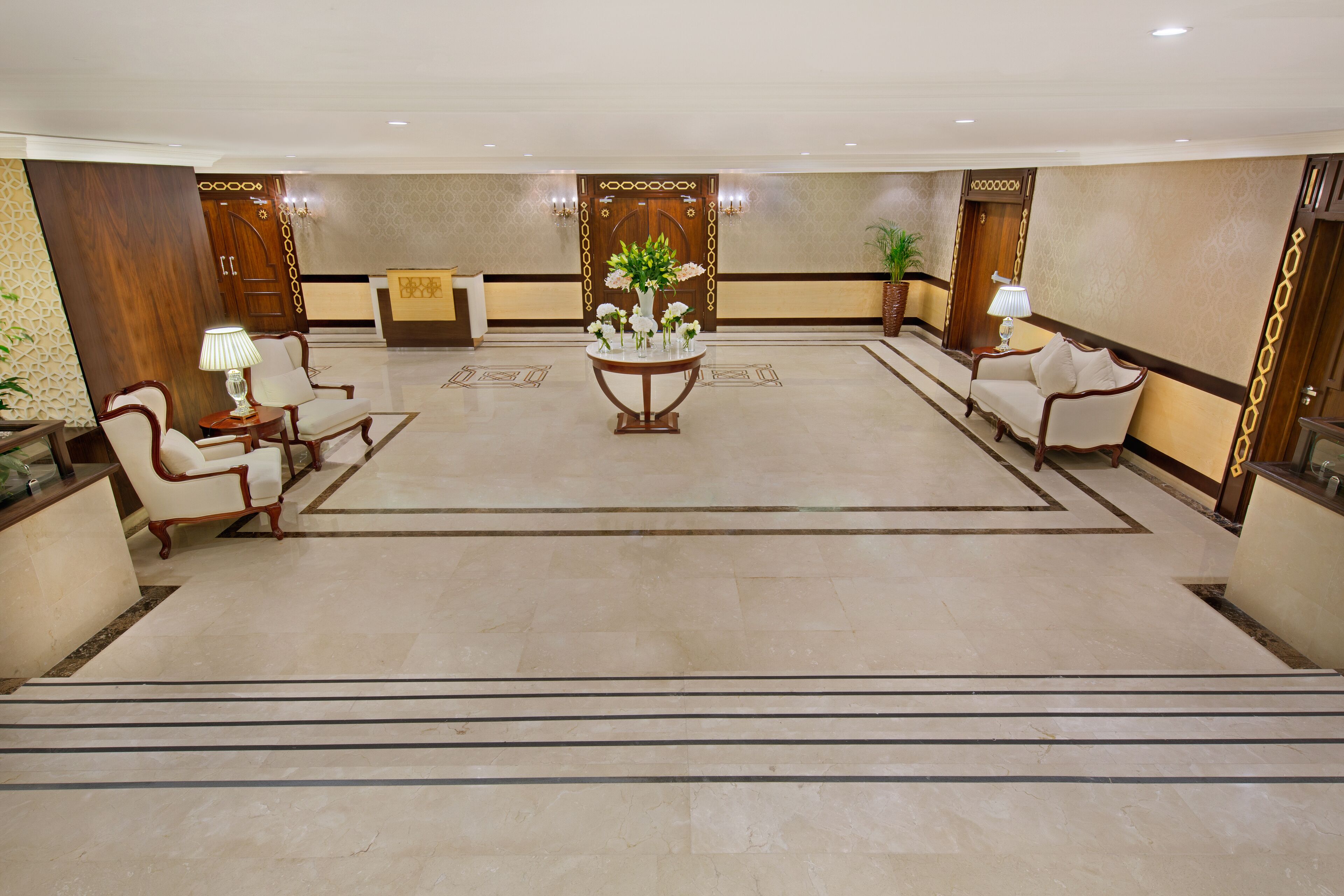 A spacious lobby with a reception desk, two sofas, a central table with a vase of flowers, and a staircase leading to upper floors.