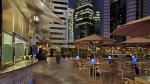 An outdoor dining area with tables and chairs, surrounded by potted plants and illuminated by ambient lighting.