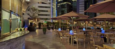 An outdoor dining area with tables and chairs, surrounded by potted plants and illuminated by ambient lighting.