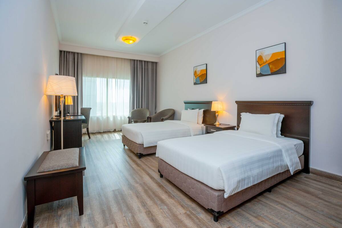 A hotel room with two beds, a desk, a chair, and a lamp.