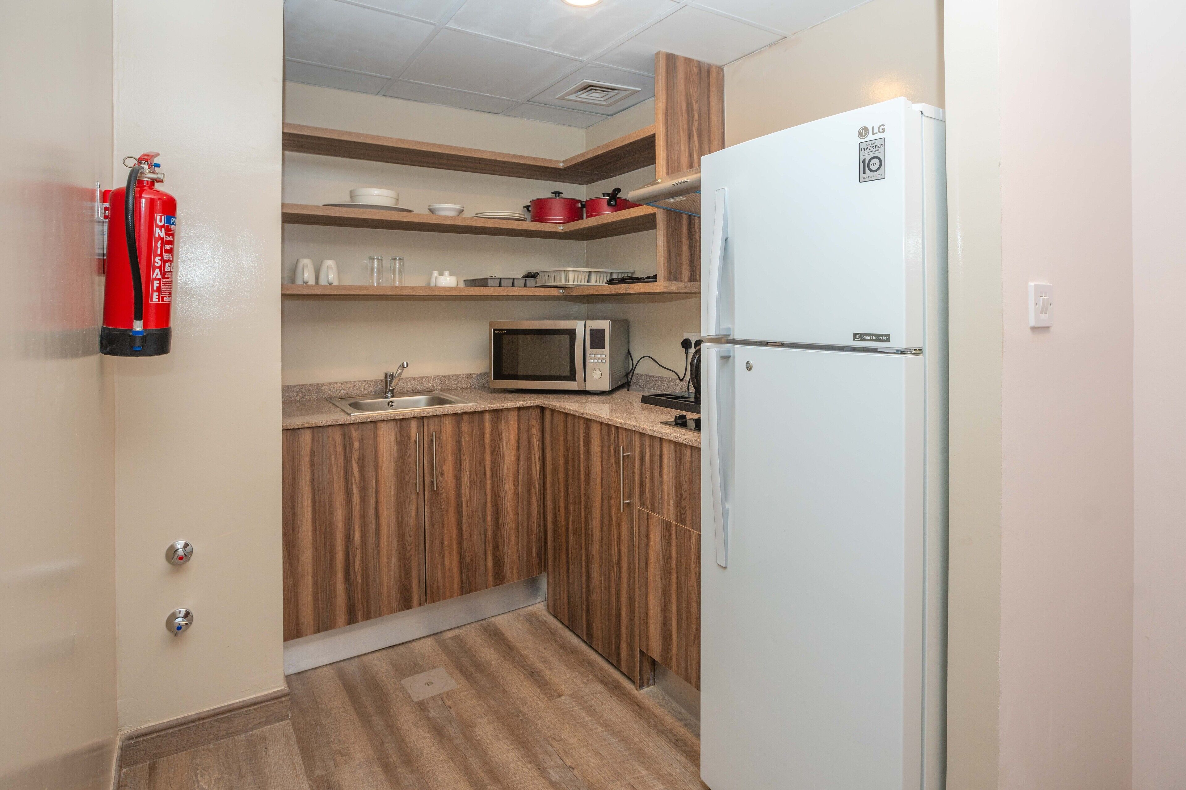 A compact kitchen with wooden cabinets, a microwave, a white refrigerator, and a fire extinguisher mounted on the wall.