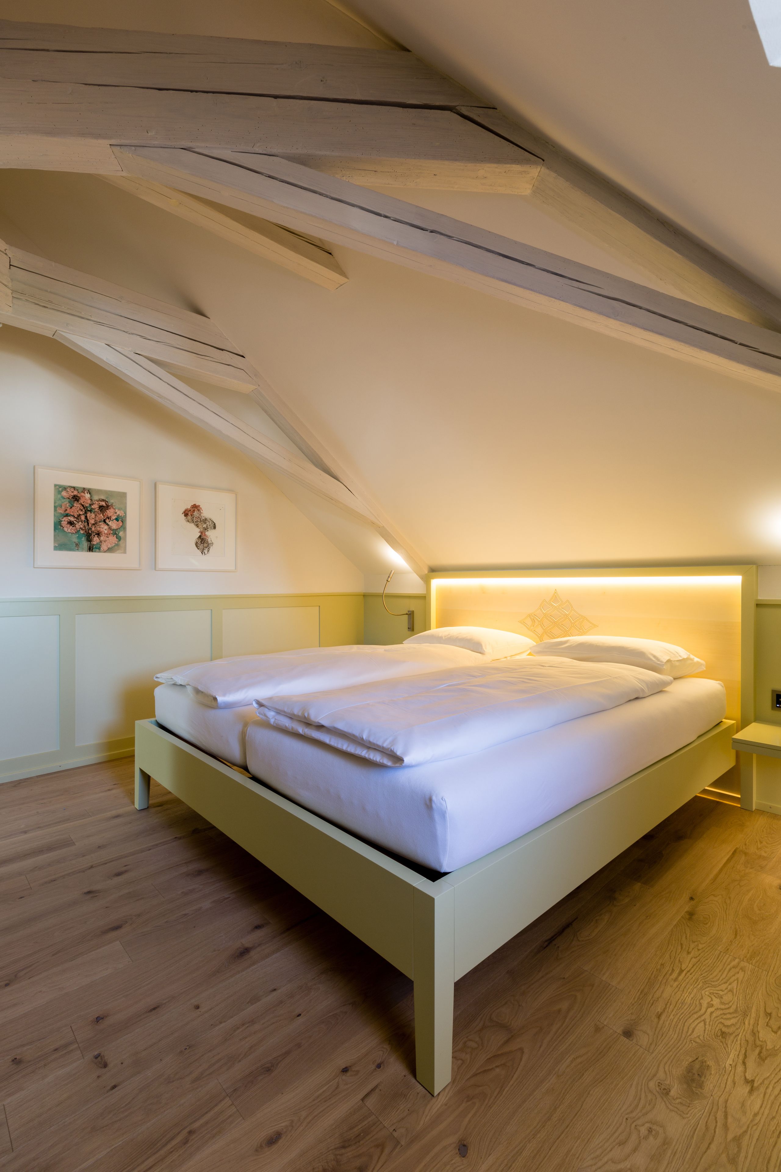 Une chambre avec un plafond en pente, un lit avec une literie blanche, un plancher en bois et deux tableaux encadrés au mur.