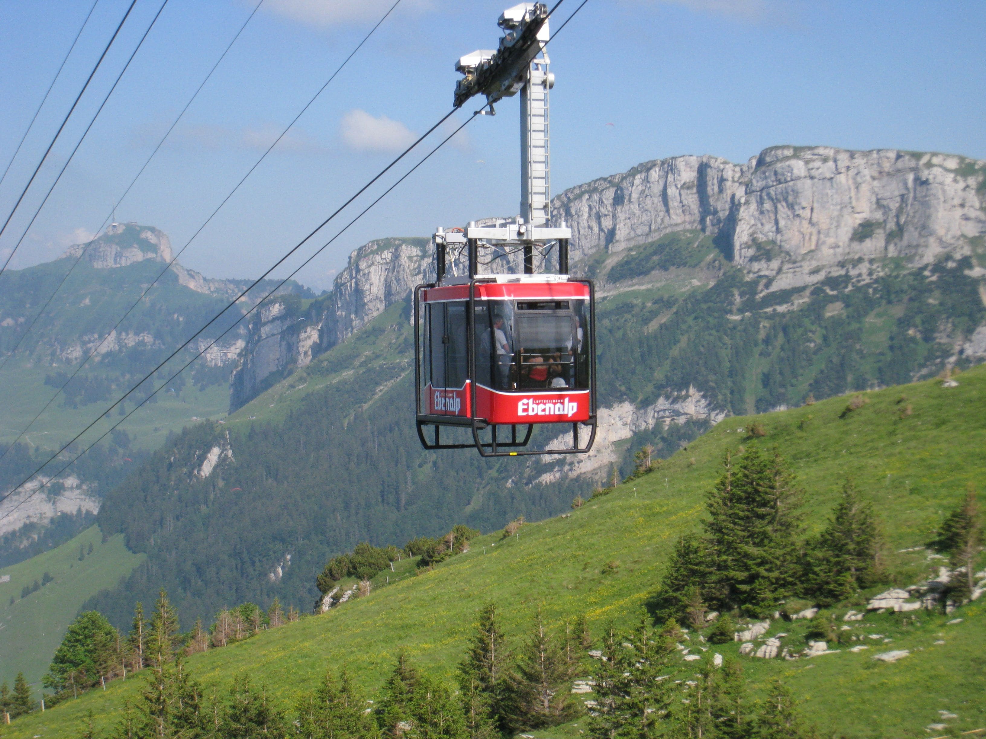 En kabelbane med 'Ebenalp' skrevet på siden, der bevæger sig langs en kabel mod en baggrund af bjerge og grøn vegetation.