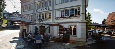 Un edificio con el nombre "KRONE" en la fachada, mesas al aire libre con sombrillas y una calle adoquinada.