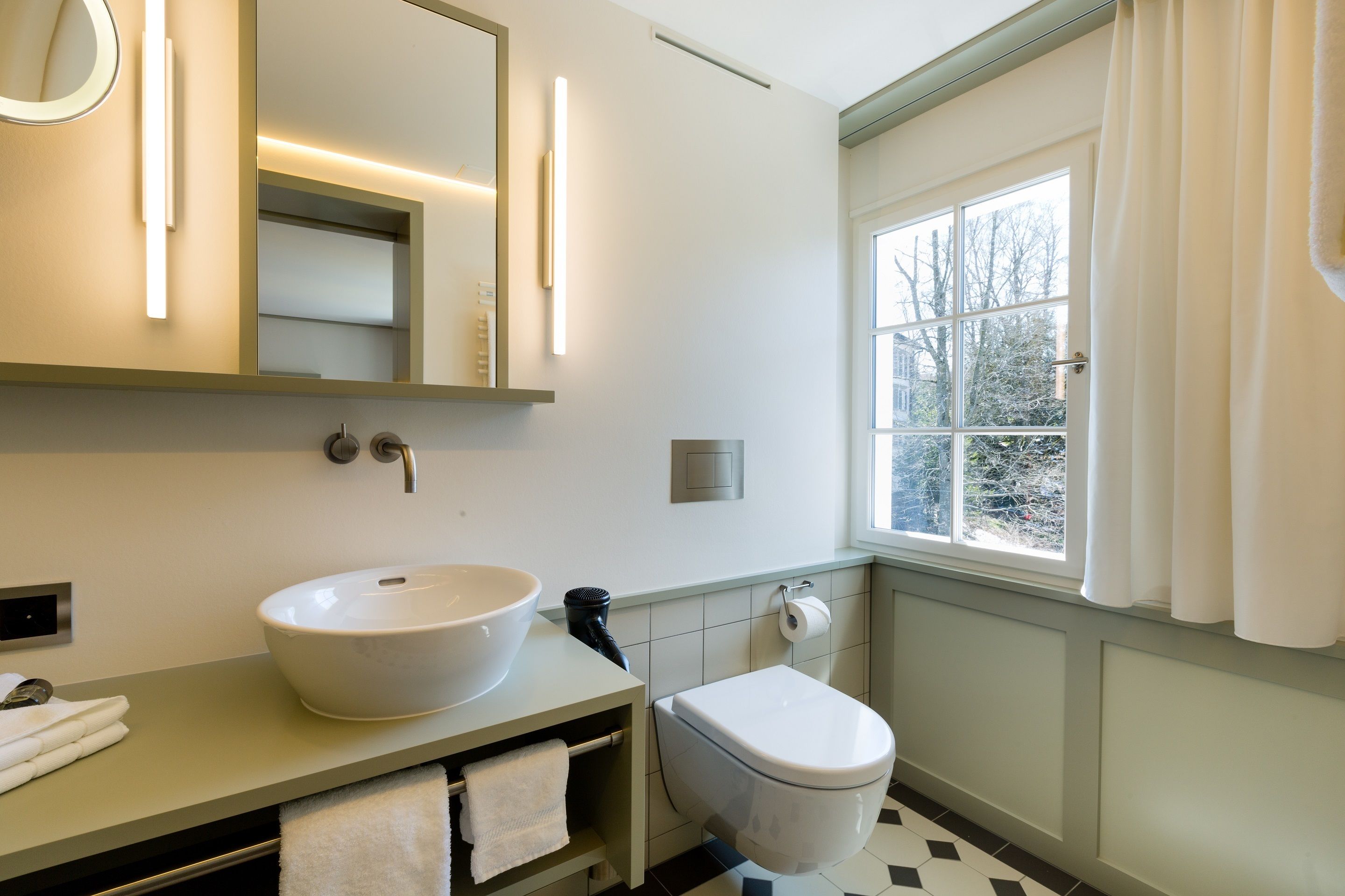 Een badkamer met een grote spiegel, een witte wastafel, een toilet en een raam met gordijnen.