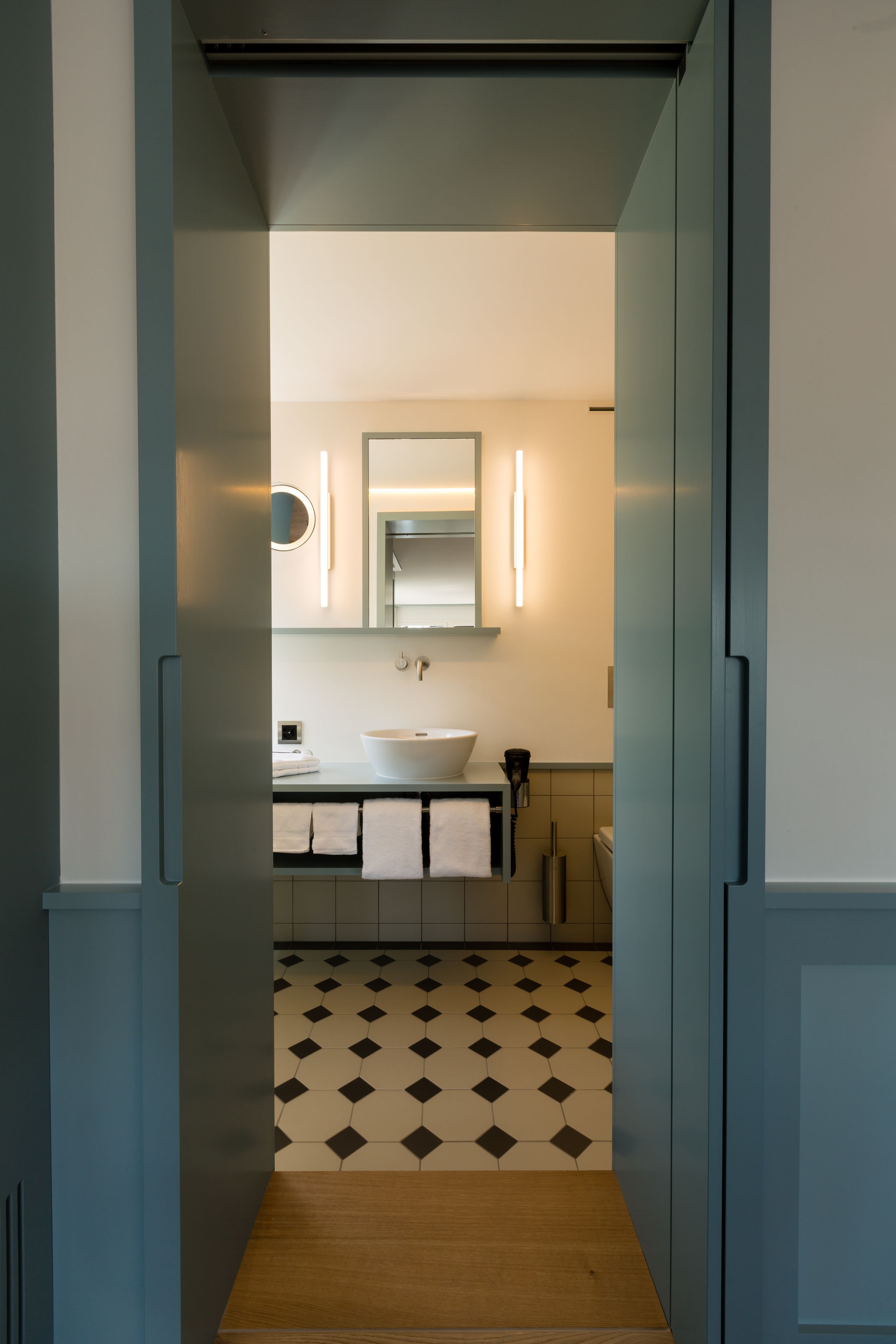A narrow hallway leading to a bathroom with a white sink, a mirror, and a patterned floor.