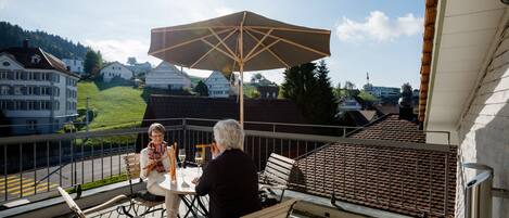 A balcony with a table and chairs, a large umbrella, and a view of houses and hills in the background.