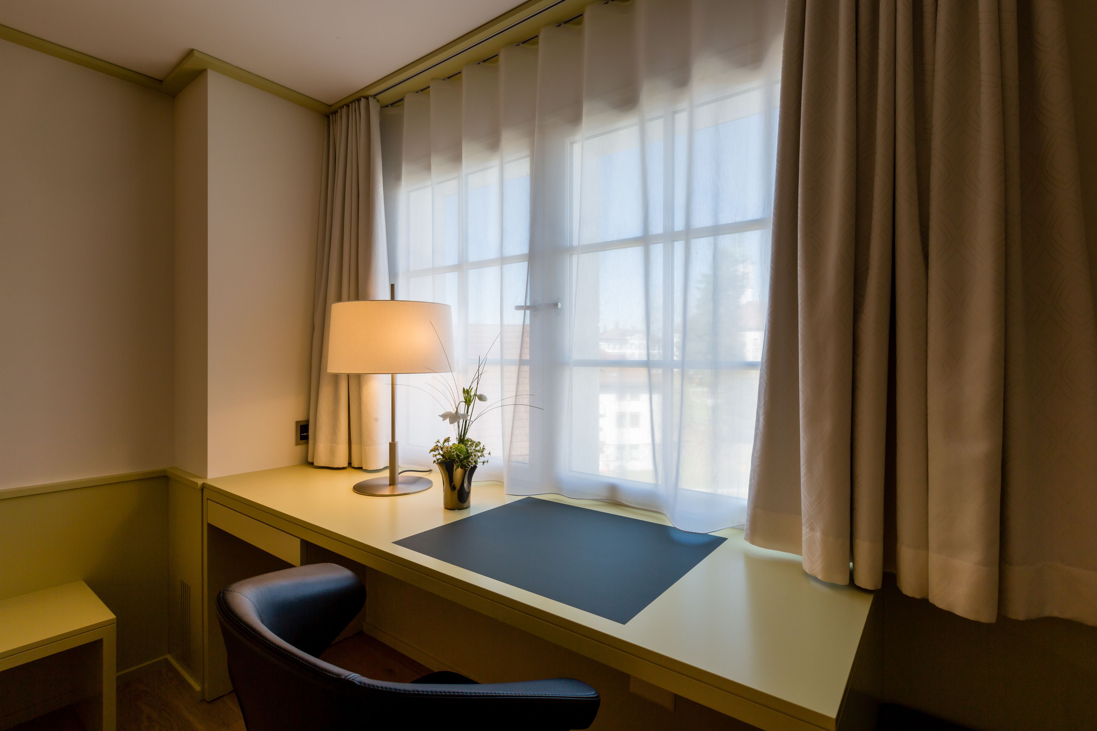 A room with a desk, chair, lamp, and window with curtains.
