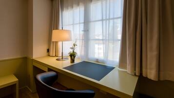 A room with a desk, chair, lamp, and window with curtains.