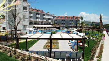 Zona de piscina con sillones de descanso y sombrillas, rodeada de un jardín ajardinado y un edificio con varios balcones.