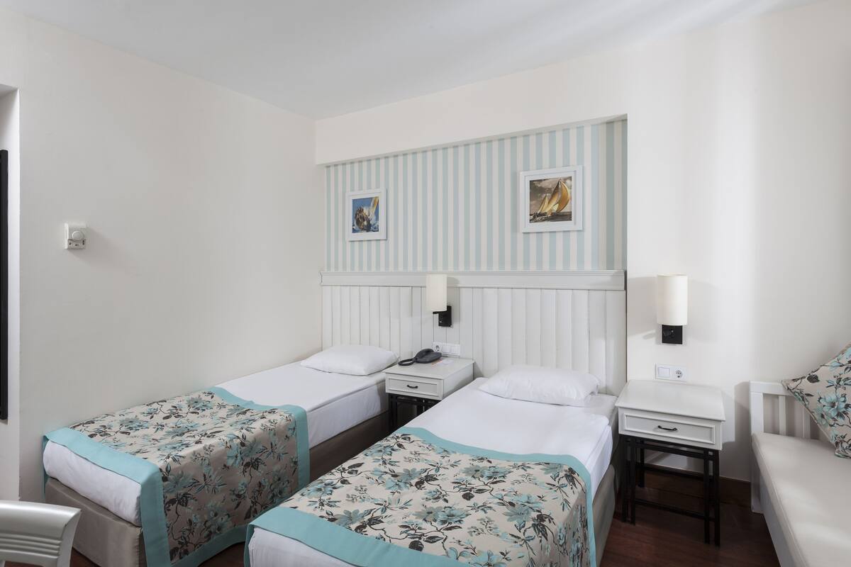 Quarto de hotel com duas camas de solteiro, uma pequena escrivaninha, uma cadeira e uma mesinha de cabeceira.